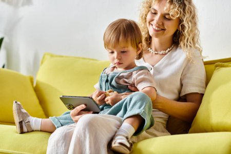 Eine lockige Mutter sitzt mit ihrer kleinen Tochter auf dem Schoß auf einer Couch und genießt einen gemütlichen Moment zu Hause.
