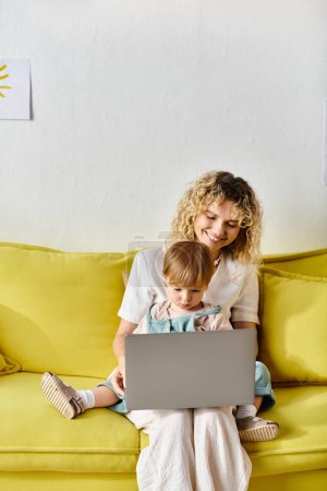 A curly-haired mother sits on a couch with her toddler daughter using a laptop together at home.
