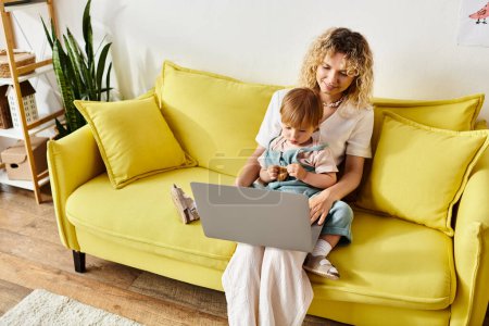 Téléchargez les photos : Une mère aux cheveux bouclés assise sur un canapé, embrassant sa petite fille dans un moment chaleureux et aimant à la maison. - en image libre de droit
