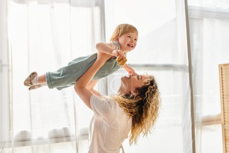 A curly mother joyfully lifts her toddler daughter up into the air, expressing love and playfulness at home.