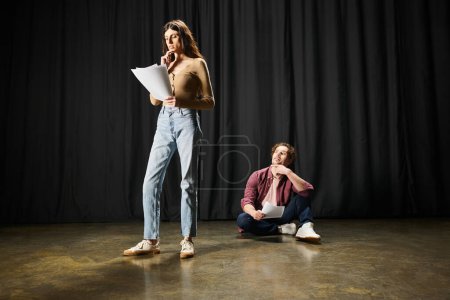 A woman and man stand confidently on a theater stage, rehearsing their roles.