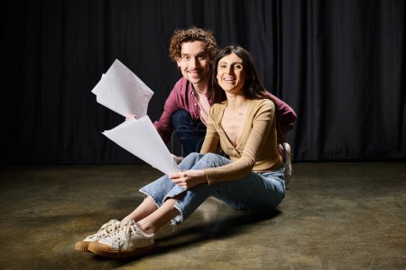 A man and woman, immersed in rehearsal, sit holding papers on the ground.