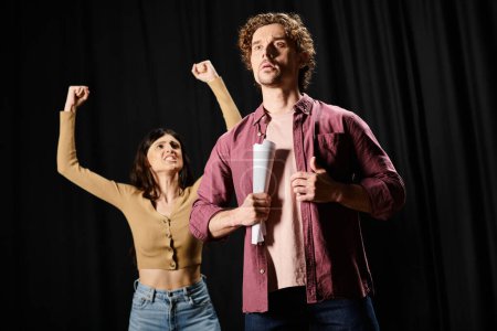 A man and a woman stand confidently in front of a microphone during rehearsals in a theater.