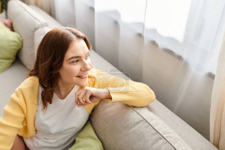 Middle aged woman in contemplation, perched on couch by a window.