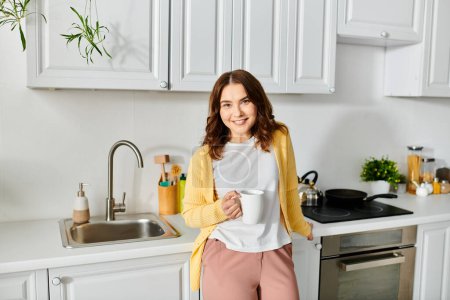 Femme d'âge moyen debout dans la cuisine, tenant une tasse.