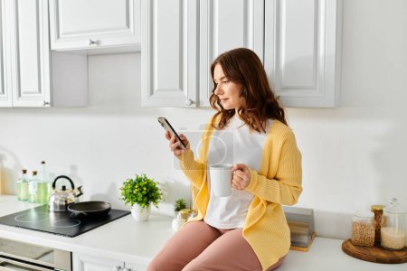 Téléchargez les photos : Une femme d'âge moyen est assise sur un comptoir de cuisine, absorbée par son téléphone portable. - en image libre de droit