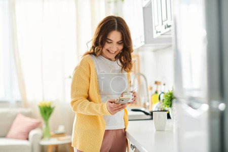 Téléchargez les photos : Une femme d'âge moyen debout dans une cuisine, concentrée sur son téléphone portable. - en image libre de droit