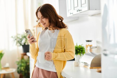 Téléchargez les photos : Une femme d'âge moyen debout dans une cuisine, absorbée dans son téléphone portable. - en image libre de droit