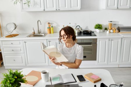 Téléchargez les photos : Une femme d'âge moyen dans un livre à sa table de cuisine. - en image libre de droit