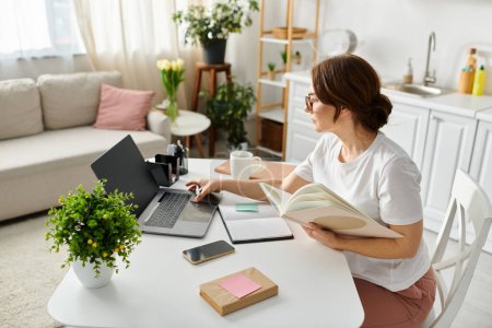Frau mittleren Alters vertieft in Arbeit, Laptop und Buch auf dem Tisch.