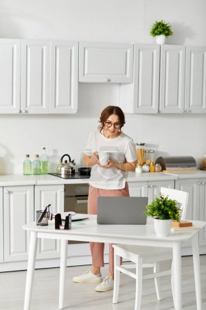 Schönheit mittleren Alters steht in der Küche, Laptop in der Hand.