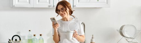 Téléchargez les photos : Femme d'âge moyen debout dans la cuisine, tenant un téléphone portable. - en image libre de droit