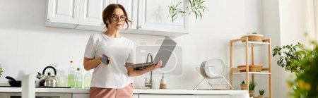 Una mujer de mediana edad se para en su cocina sosteniendo un portátil, mezclando tecnología y cocinando.