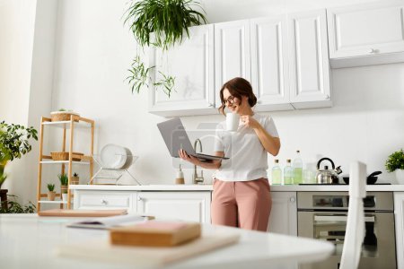 Middle-aged woman standing in kitchen, multitasking with laptop in hand.