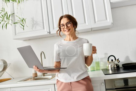 A middle-aged woman balancing a laptop and coffee cup at home.