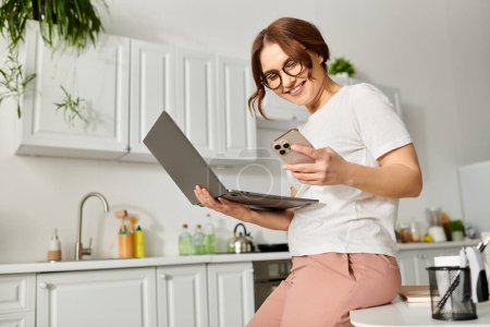 Photo for Middle-aged woman sits on counter, typing on laptop. - Royalty Free Image