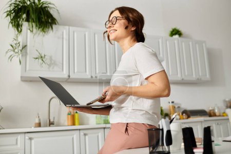Femme d'âge moyen multitâche dans la cuisine, tenant un ordinateur portable.