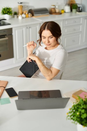 Middle aged woman sitting at kitchen table working on laptop.