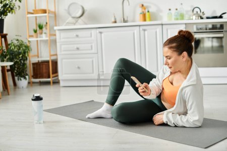 Femme d'âge moyen assise sur un tapis de yoga, absorbée par le téléphone.