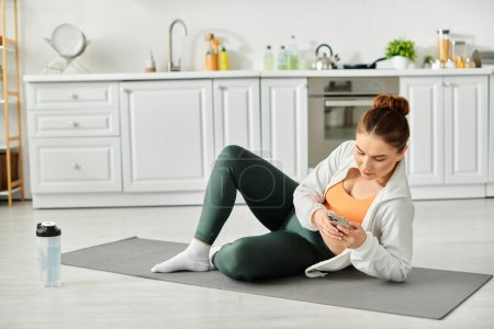 Middle-aged woman seated on yoga mat, captivated by smartphone screen.
