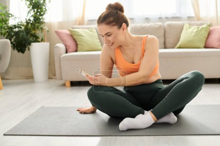 Téléchargez les photos : Femme d'âge moyen est assis sur tapis de yoga, absorbé dans le téléphone. - en image libre de droit