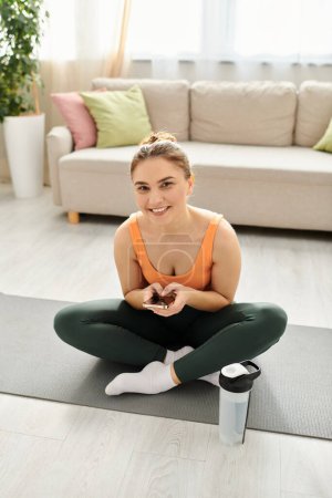 Femme d'âge moyen assis sur tapis de yoga avec téléphone.