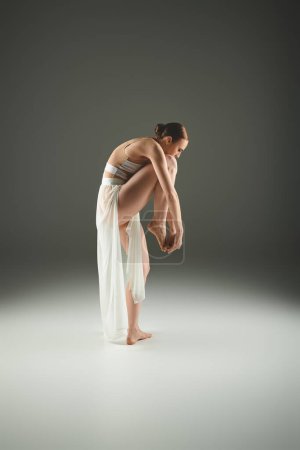 A young, beautiful ballerina in a white dress gracefully bends over.