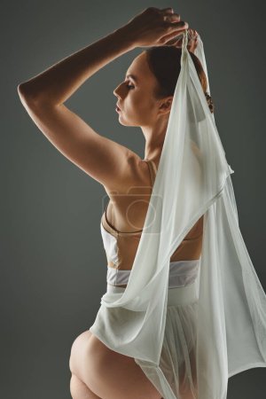 A young woman in a white dress gracefully holds a white scarf over her head.