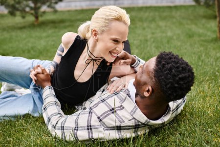 Foto de Una feliz pareja multicultural, un hombre afroamericano y una mujer caucásica, relajándose juntos en la exuberante hierba verde de un parque. - Imagen libre de derechos