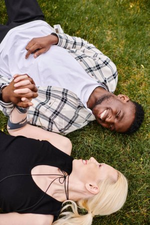 Un hombre afroamericano y una mujer caucásica yacen en la hierba, abrazándose con sonrisas en sus rostros.