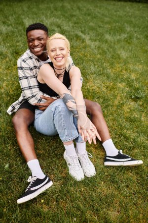 Una pareja multicultural, un hombre afroamericano y una mujer caucásica, sentados contentos en la hierba de un parque.