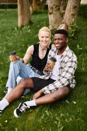 Foto de Una pareja feliz y multicultural, un hombre afroamericano y una mujer caucásica, sentados juntos en el césped en un parque. - Imagen libre de derechos