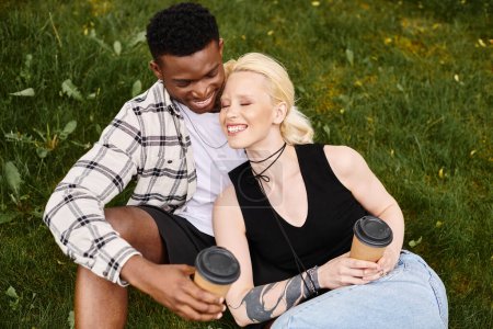 Foto de Una pareja feliz y multicultural - un hombre afroamericano y una mujer caucásica - sentados juntos en el césped en un parque. - Imagen libre de derechos