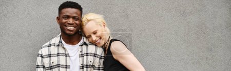 Téléchargez les photos : Un homme et une femme heureux et multiculturels côte à côte, embrassant une rue urbaine près d'un bâtiment gris. - en image libre de droit