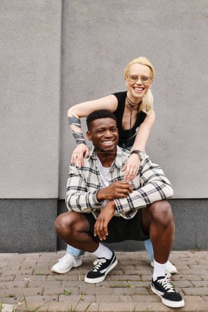Un alegre novio multicultural y novia posan juntos en una calle urbana cerca de un edificio gris, sonriendo a la cámara.
