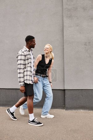 Una feliz pareja multicultural, novio y novia, caminando juntos en una calle urbana cerca de un edificio gris.