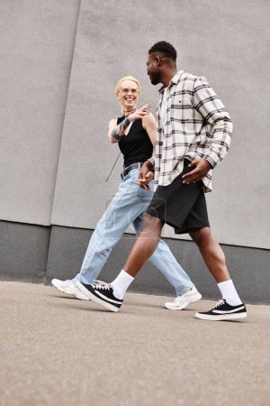 Foto de Una feliz pareja multicultural camina por una calle urbana junto a un edificio gris, disfrutando de la compañía de los demás. - Imagen libre de derechos
