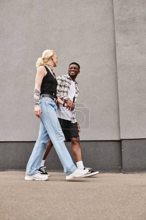 Una pareja multicultural, un hombre y una mujer, caminando felizmente por una calle urbana cerca de un edificio gris.
