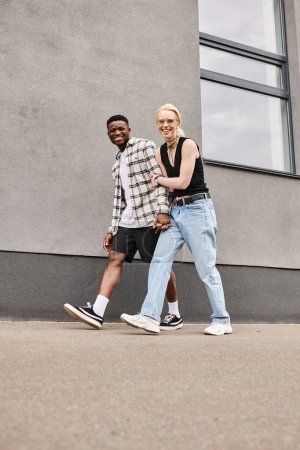 A happy multicultural boyfriend and girlfriend walk together on an urban street near a grey building.