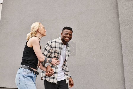 A happy couple, a multicultural boyfriend and girlfriend, walk together on an urban street near a grey building.
