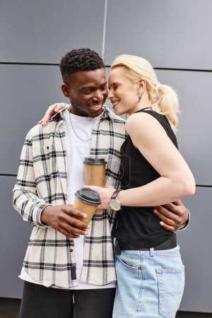 A multicultural couple, a man, and a woman, hug each other while holding coffee cups on an urban street near a grey building.