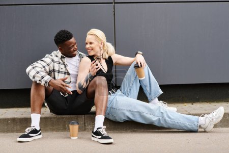 Téléchargez les photos : Un homme et une femme de différentes ethnies regardent le contenu alors qu'ils sont assis côte à côte sur le sol près d'un bâtiment gris dans la ville. - en image libre de droit