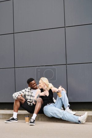 A happy multicultural couple sitting beside each other on the ground near a grey urban building, sharing a quiet moment.