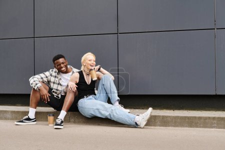 A happy multicultural couple sitting beside each other on the ground near a grey urban building
