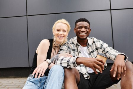 Téléchargez les photos : Un heureux couple multiculturel assis sur le sol près d'un bâtiment gris dans une rue urbaine, partageant un moment de convivialité. - en image libre de droit
