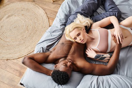 Un novio y una novia multiculturales están tendidos románticamente entrelazados en una cama en casa, compartiendo un momento de intimidad juntos.