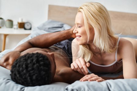 A sensual moment captured as a multicultural man and woman embrace intimately while laying on a bed at home.