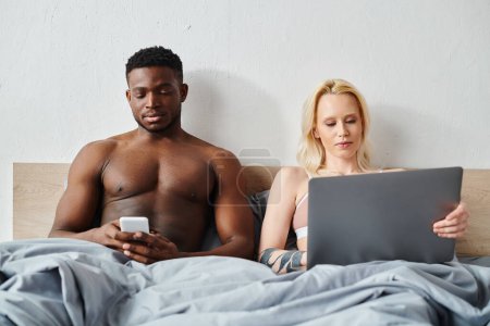A multicultural boyfriend and girlfriend sit on a bed, focused on their devices.