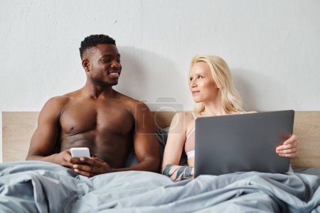 Téléchargez les photos : Un homme et une femme multiculturels assis sur un lit, absorbés par l'écran d'un ordinateur portable devant eux. - en image libre de droit