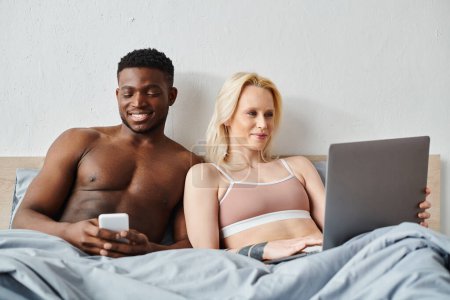 A multicultural couple sits on a bed, engrossed in a laptop screen together.